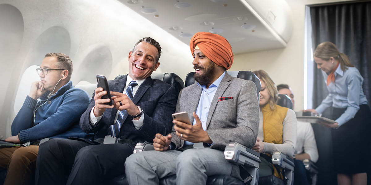 A Caucasian man and a Sikh man sitting next to each other on a plane. They happily converse with each other.