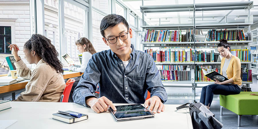 A young man in a library, reading the brochure “Was Life Created?” on his tablet.