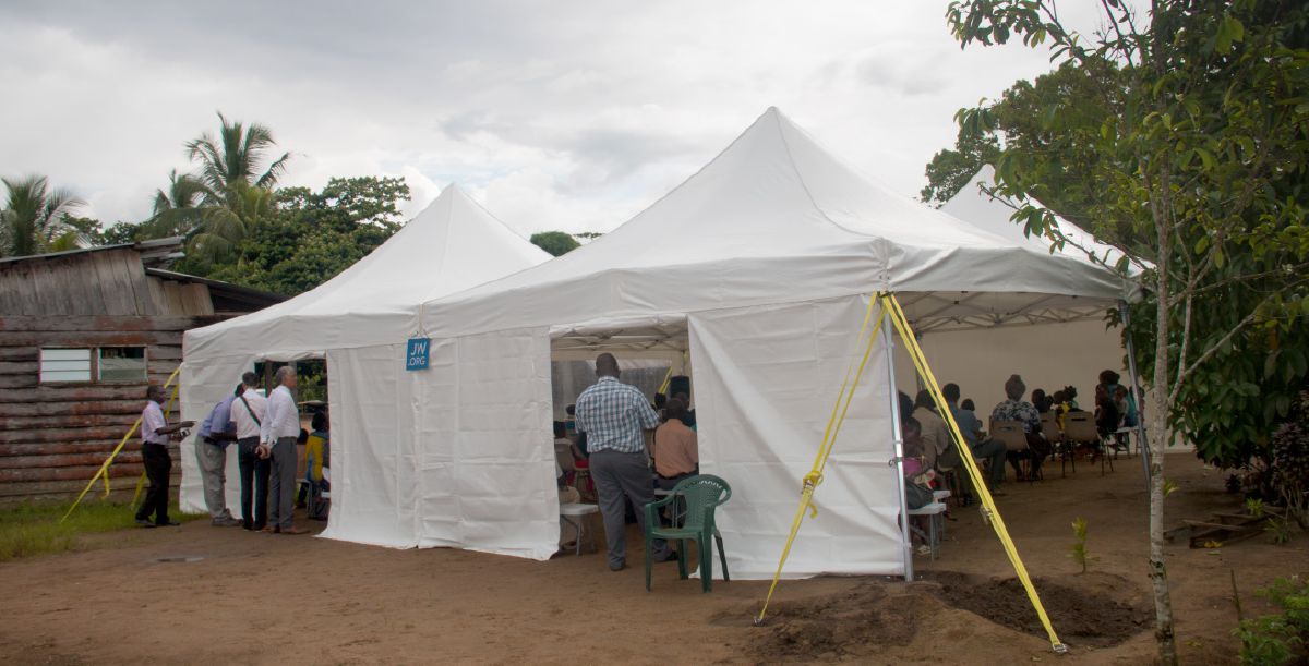A group of people attending a public meeting being held in a large tent set up in the forest.