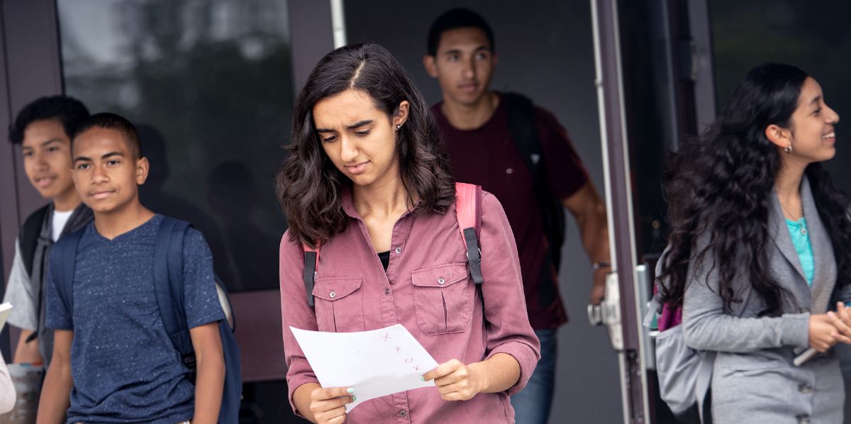 Several teenagers exiting a school. One of the girls is looking disappointedly at her exam results.