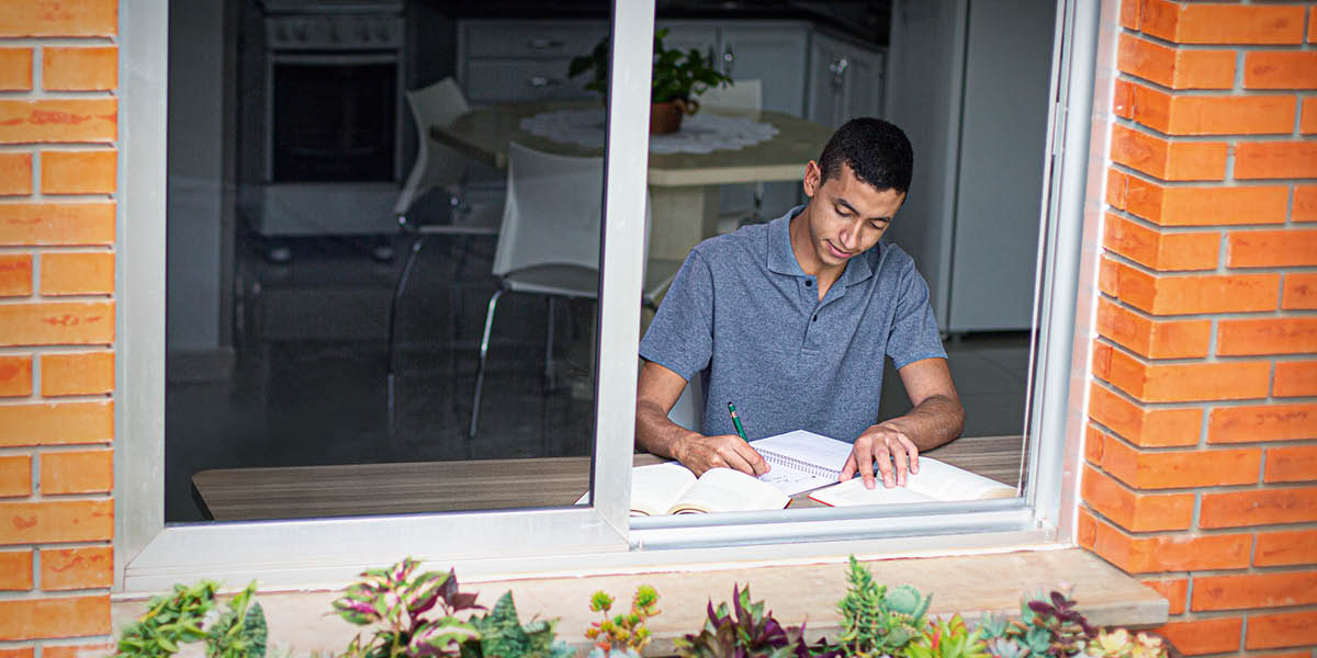 A teenage boy studying at a table near a window of his home.