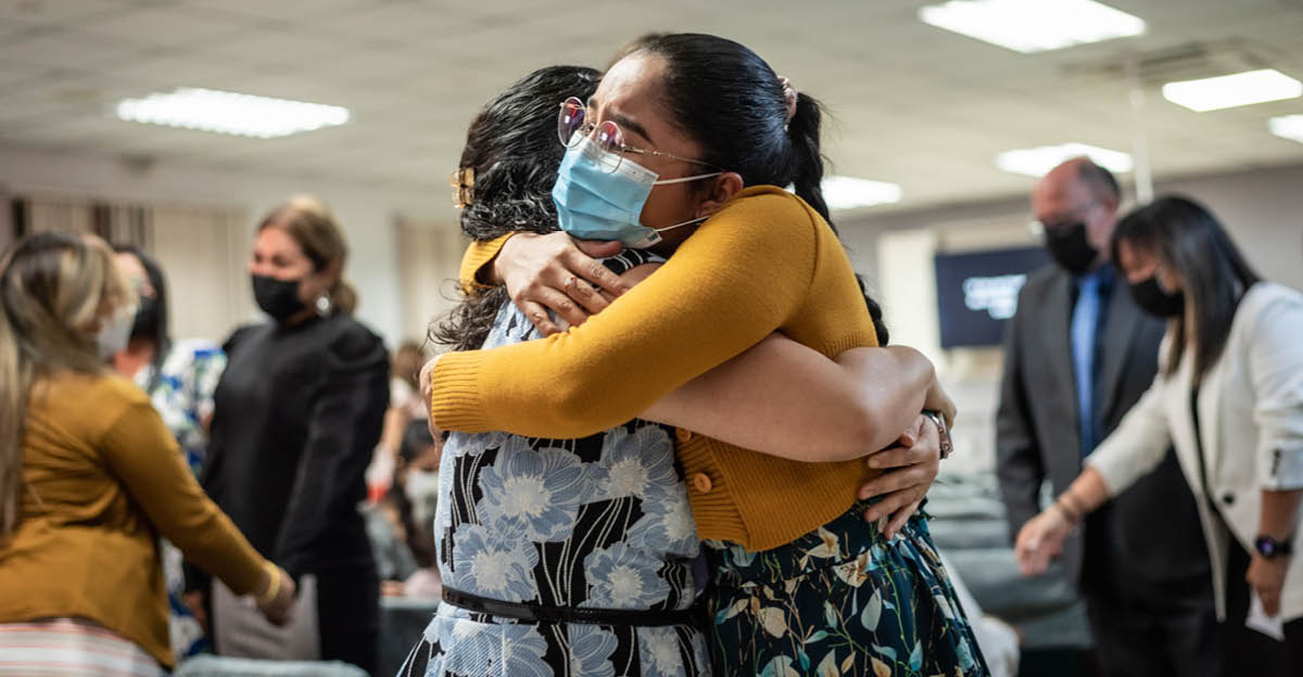 Two sisters wearing masks, hugging at a Kingdom Hall. In the background, brothers and sisters wearing masks greet each other warmly.