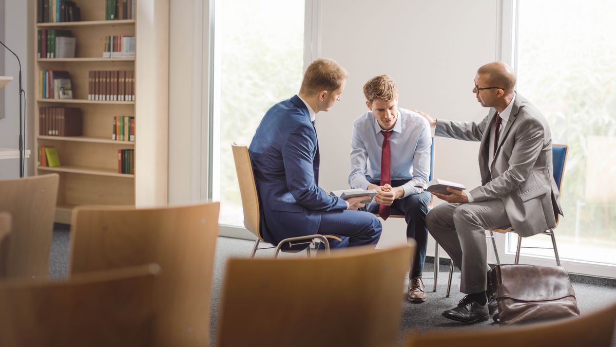 Two congregation elders kindly sharing Bible verses with a young man. One of the elders rests his hand on the young man’s shoulder as he speaks to him.