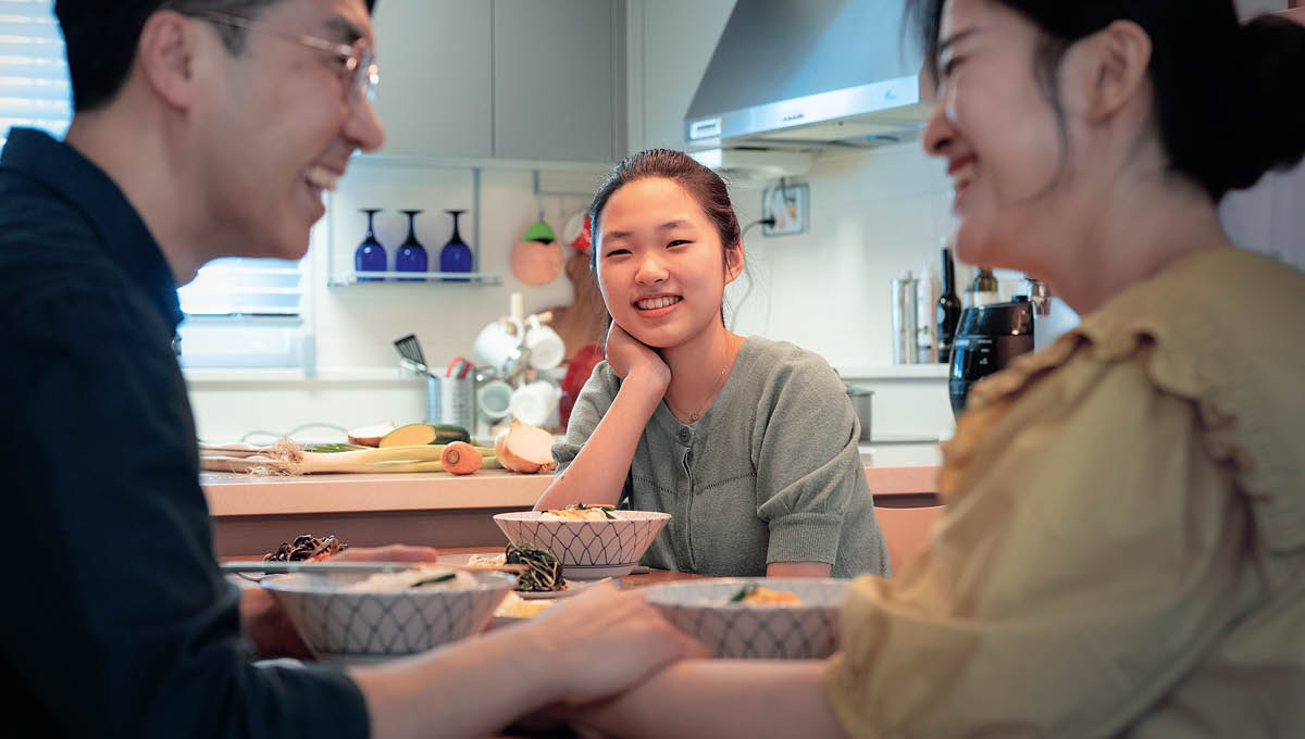 A teenage daughter happily observing her parents at the dinner table. Her father holds her mother’s hand as they laugh together.