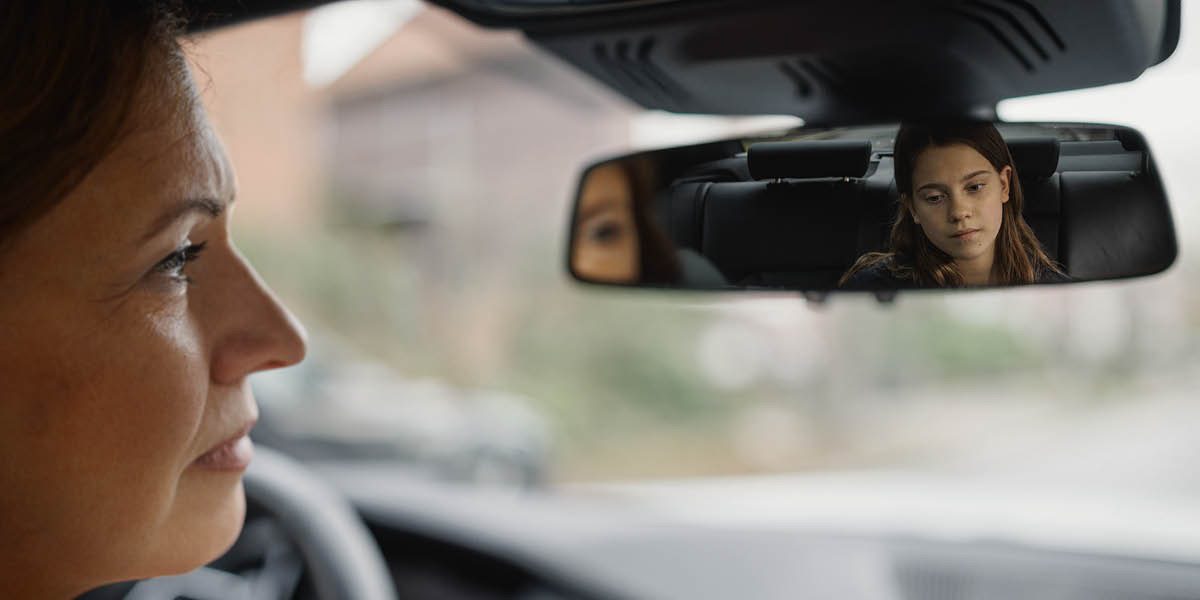 A mother observing in her car’s rearview mirror the gloomy expression on her daughter’s face.