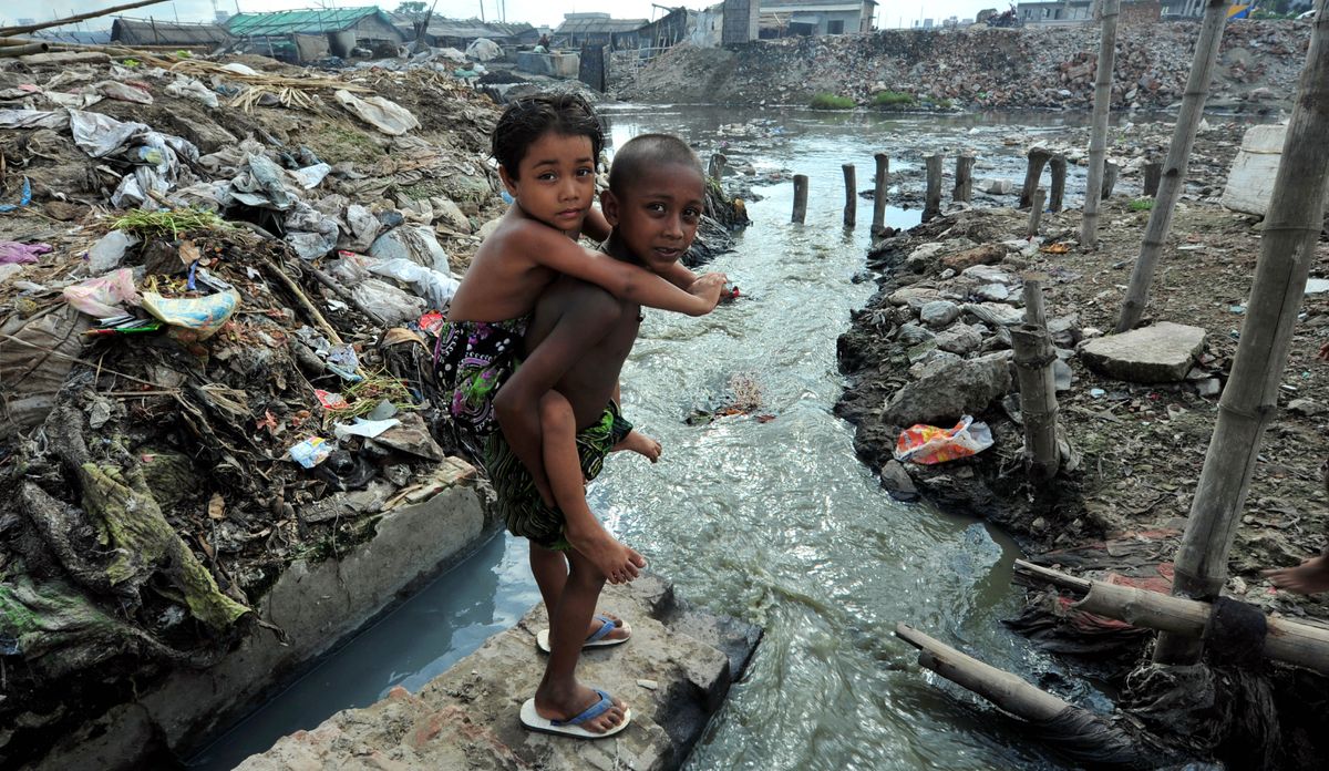 An older boy carrying a younger boy on his back at the edge of a heavily polluted stream.