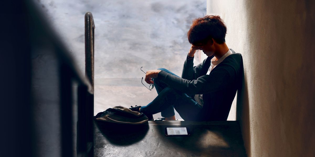 A teenager resting his head in his hand as he sits alone at the bottom of a set of stairs in a dark hallway.